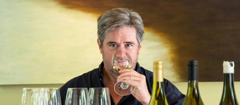 Food and wine executive wine editor Ray Isle sniffing a glass of wine while sitting behind a table full of wine glasses and three opened bottles of wine. Photo credit to Mark Fiorito